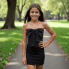 A young girl wearing a black strapless dress with a bow at the chest, standing in a park setting.