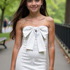 A woman wearing a white mini dress with a bow detail on the chest, standing in a park setting.