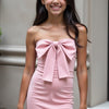 A woman standing outside wearing a pink strapless mini dress with a bow detail at the bust.